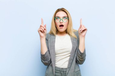 Şok olmuş, şaşkın ve ağzı açık genç, sarışın bir kadın, iki eliyle yukarıya bakıyor düz duvara uzayı kopyalamak için