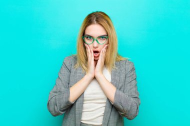 Genç, güzel, sarışın bir kadın şok olmuş ve korkmuş hissediyor. Ağzı açık ve elleri düz duvarda.