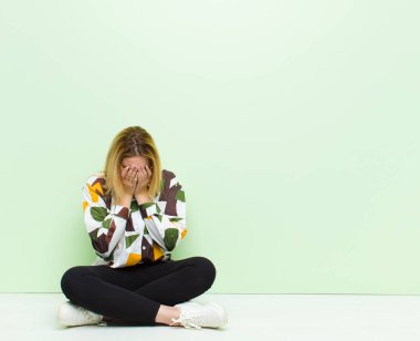 Genç sarışın kadın üzgün, hayal kırıklığına uğramış, gergin ve depresif, iki eliyle yüzünü kapatmış, yerde oturmuş ağlıyor.