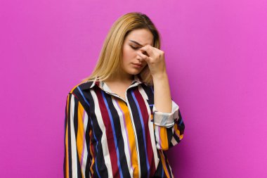 Genç sarışın kadın stresli, mutsuz ve hayal kırıklığına uğramış, alnına dokunuyor ve mor duvara karşı şiddetli baş ağrısı çekiyor.
