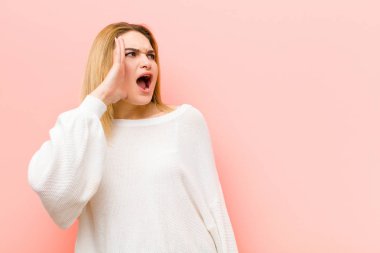 Genç, güzel sarışın kadın öfkeyle ve yüksek sesle bağırıyor kenardaki boşluğu kopyalamak için, eli ağızının yanında pembe düz duvara karşı.
