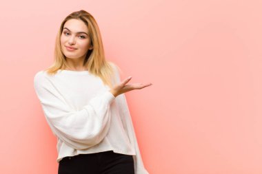 Genç, güzel, sarışın bir kadın neşeyle gülümsüyor, mutlu hissediyor ve fotokopi odasında bir konsept gösteriyor. Avuç içi pembe düz duvara dayalı.