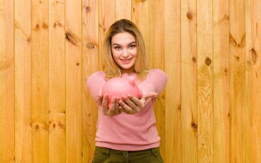 Genç, güzel, sarışın bir kadın, kumbarası ahşap duvara dayalı.