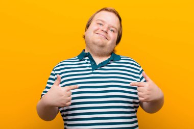 Genç, cüsseli, gururlu, pozitif ve rahat görünen, iki elini de turuncu duvara dayamış göğsünü gösteren bir adam.