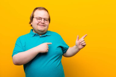 Genç, iri yarı, şaşkın ve şaşırmış hisseden adam, yan taraftaki fotokopi alanını işaret ederek turuncu duvara karşı şaşkın, açık ağızlı bir bakış atıyor.