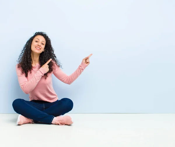 young pretty woman smiling happily and pointing to side and upwards with both hands showing object in copy space sitting on floor