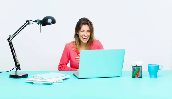 young pretty woman working with a laptop laughing out loud at some hilarious joke, feeling happy and cheerful, having fun
