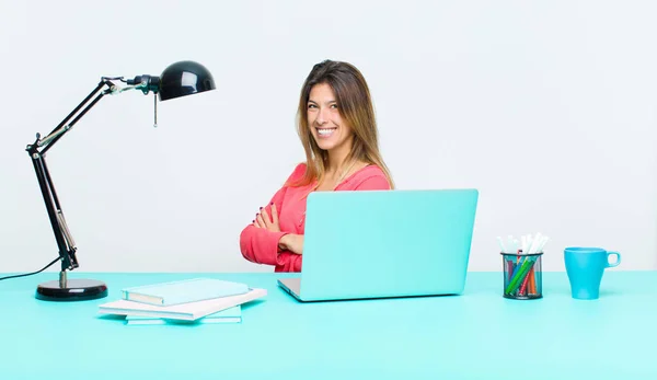 young pretty woman working with a laptop smiling to camera with crossed arms and a happy, confident, satisfied expression, lateral view