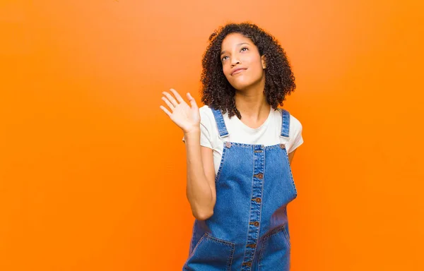 young pretty black woman smiling happily and cheerfully, waving hand, welcoming and greeting you, or saying goodbye against orange wall