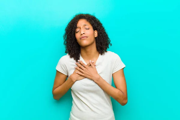 young black woman looking sad, hurt and heartbroken, holding both hands close to heart, crying and feeling depressed against blue wall