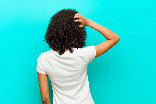 young black woman thinking or doubting, scratching head, feeling puzzled and confused, back or rear view against blue wall