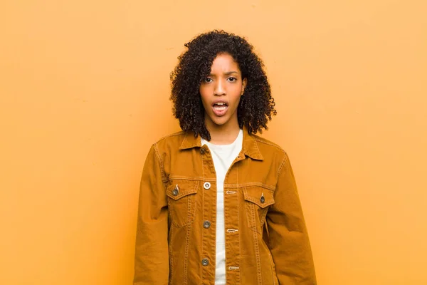 young pretty black woman looking very shocked or surprised, staring with open mouth saying wow against orange wall