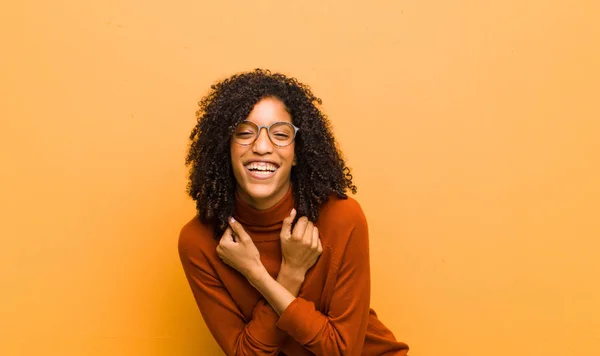 young pretty black woman smiling cheerfully and celebrating, with fists clenched and arms crossed, feeling happy and positive against orange wall