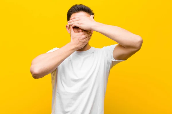 young handsome man covering face with both hands saying no to the camera! refusing pictures or forbidding photos against orange wall