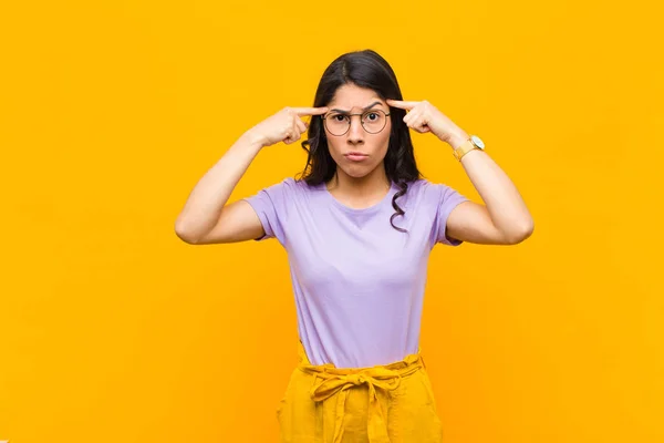 young pretty latin woman with a serious and concentrated look, brainstorming and thinking about a challenging problem against orange wall