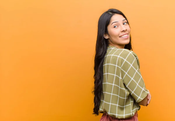 young pretty hispanic woman smiling gleefully, feeling happy, satisfied and relaxed, with crossed arms and looking to the side against brown wall