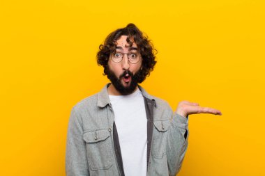 Genç çılgın adam şaşırmış ve şok olmuş görünüyor. Çenesi düşmüş. Sarı duvara karşı eli açık bir obje tutuyor.