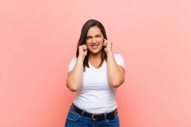 Genç, güzel İspanyol bir kadın kızgın, stresli ve sinirli görünüyor, her iki kulağı da sağır edici bir ses, ses veya pembe duvarın karşısında yüksek sesle müzik dinliyor.