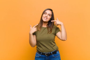 Genç, güzel bir kadın, kendine güvenerek gülümsüyor. Kendi gülüşünü gösteriyor. Olumlu, rahat, tatmin olmuş, turuncu duvara yaslanmış.