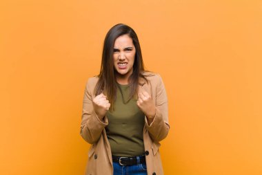 Genç ve güzel bir kadın sinirli sinirli, sinirli bakışlar ve sıkı yumruklarla bağırıyor. Turuncu duvara karşı öfkeli bir şekilde izole edilmiş hissediyor.