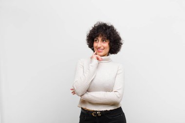 Genç ve güzel afro kadın mutlu bir şekilde gülümsüyor ve hayal kuruyor ya da şüphe ediyor, yan tarafa bakıyor.