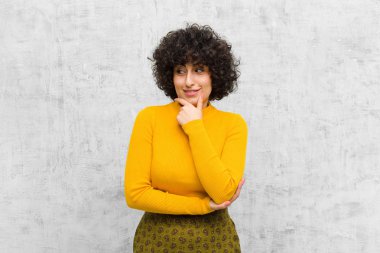 Genç, güzel afro bir kadın mutlu, kendinden emin bir ifadeyle gülümsüyor, çenesinde elleri, merak ediyor ve yan tarafa bakıyor.