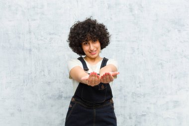 Genç, güzel afro kadın mutlu bir şekilde gülümsüyor, arkadaş canlısı, kendinden emin, pozitif bakışlı, bir nesne ya da konsept sunuyor ve gösteriyor.