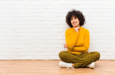 Genç, güzel afro kadın mutlu bir şekilde gülümsüyor ve hayal kuruyor ya da şüpheyle bakıyor, yere bakıyor.