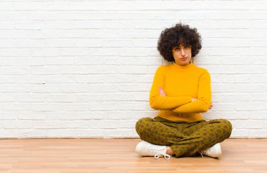 Genç, güzel, afro bir kadın hayal kırıklığına uğramış ve hayal kırıklığına uğramış, ciddi görünüyor, kızgın ve kollarını kavuşturup yerde oturuyor.