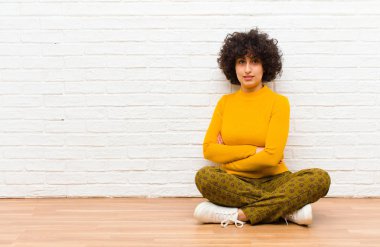 Genç, güzel afro kadın kameraya çapraz kollarıyla gülümsüyor ve mutlu, kendinden emin, tatmin olmuş bir ifadeyle yerde yan yana oturuyor.