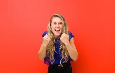 Genç sarışın kadın zaferle bağırıyor, gülüyor, mutlu ve heyecanlı kırmızı duvara karşı başarıyı kutlarken.