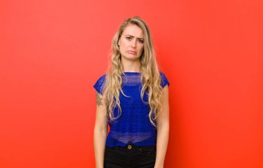 Genç, sarışın bir kadın üzgün ve mızmız görünüyor. Mutsuz görünüyor. Kırmızı duvara karşı olumsuz ve hayal kırıklığına uğramış bir tavırla ağlıyor.