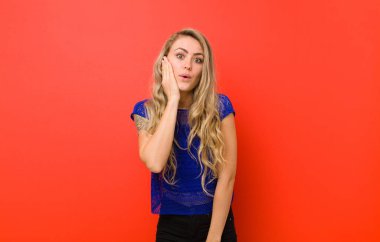 Genç sarışın kadın şok olmuş ve şaşkınlık içinde. Kırmızı duvara karşı ağzı sonuna kadar açık bir şekilde inanmayarak el ele tutuşuyor.