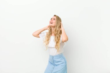 Genç sarışın kadın gülümsüyor ve rahatlıyor, tatmin oluyor ve kaygısız, pozitif gülüyor ve beyaz duvara yaslanıyor.