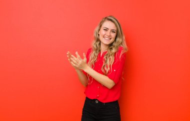 Genç sarışın kadın gülümsüyor ve arkadaş canlısı görünüyor. İki ya da iki numarayı gösteriyor. Eli önde, kırmızı duvara doğru sayıyor.