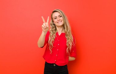 Genç sarışın kadın gülümsüyor ve mutlu görünüyor, kaygısız ve pozitif, kırmızı duvara karşı zafer ya da barış işareti yapıyor.