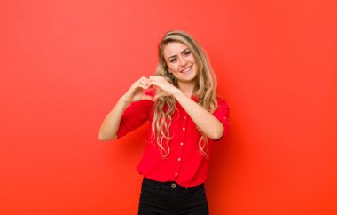 Genç sarışın kadın gülümsüyor ve mutlu, sevimli, romantik ve aşık, iki elini de kırmızı duvara dayayıp kalbini şekillendiriyor.