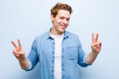 Genç kızıl saçlı adam gülümsüyor ve mutlu görünüyor, arkadaş canlısı ve memnun, mavi duvara karşı zafer ya da barış işareti yapıyor.
