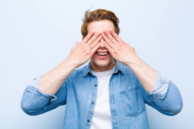 Genç kızıl saçlı adam gülümsüyor ve mutlu hissediyor, iki eliyle gözlerini kapatıyor ve mavi duvara karşı inanılmaz bir sürpriz bekliyor.