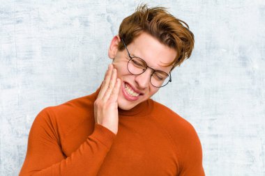 Genç kızıl saçlı, yanak tutan ve acı çeken diş ağrısı çeken, hasta, sefil ve mutsuz hisseden, grunge duvarına karşı bir dişçi arayan adam.