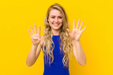 Genç sarışın kadın gülümsüyor ve arkadaş canlısı görünüyor. 9 ya da 9 numarayı gösteriyor. Eli önde, sarı duvara doğru sayıyor.