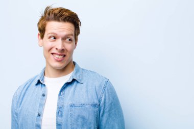 Genç kızıl saçlı adam endişeli, stresli, endişeli ve korkmuş görünüyor, panikliyor ve mavi duvara karşı dişlerini sıkıyor