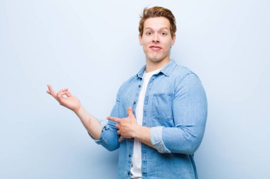 Genç kızıl saçlı adam gururla ve güvenle gülümsüyor, mutlu ve tatmin olmuş hissediyor ve mavi duvara karşı fotokopi alanında bir konsept gösteriyor.