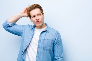 Genç kızıl saçlı adam şaşkın ve kafası karışmış hissediyor. Kafasını kaşıyor ve mavi duvara bakıyor.