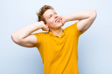 Genç kızıl saçlı adam gülümsüyor ve rahatlıyor, memnun ve kaygısız, pozitif gülüyor ve mavi duvara karşı ürperiyor.