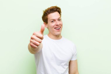 Genç kızıl saçlı yakışıklı adam gururlu, tasasız, kendinden emin ve mutlu, başparmağıyla olumlu gülümsüyor.