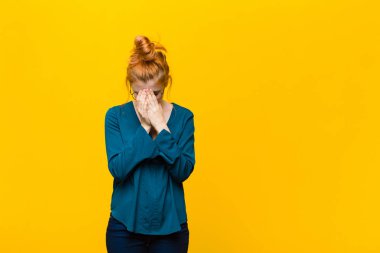 Genç kızıl saçlı kadın üzgün, sinirli ve depresif hissediyor. İki eliyle yüzünü kapatıyor. Turuncu duvara karşı ağlıyor.