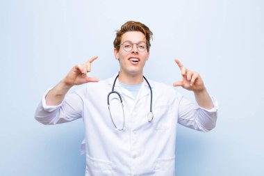Genç kızıl kafalı doktor iki eliyle kendi gülümsemesini çiziyor, olumlu ve mutlu görünüyor, mavi duvara karşı sağIık kavramı