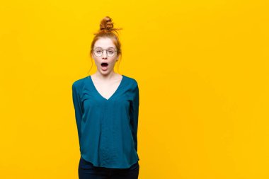 Genç kızıl saçlı kadın çok şaşırmış ya da şaşkın görünüyor, ağzı açık turuncu duvara karşı 