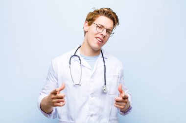 young red head doctor smiling with a positive, successful, happy attitude pointing to the camera, making gun sign with hands against blue wall